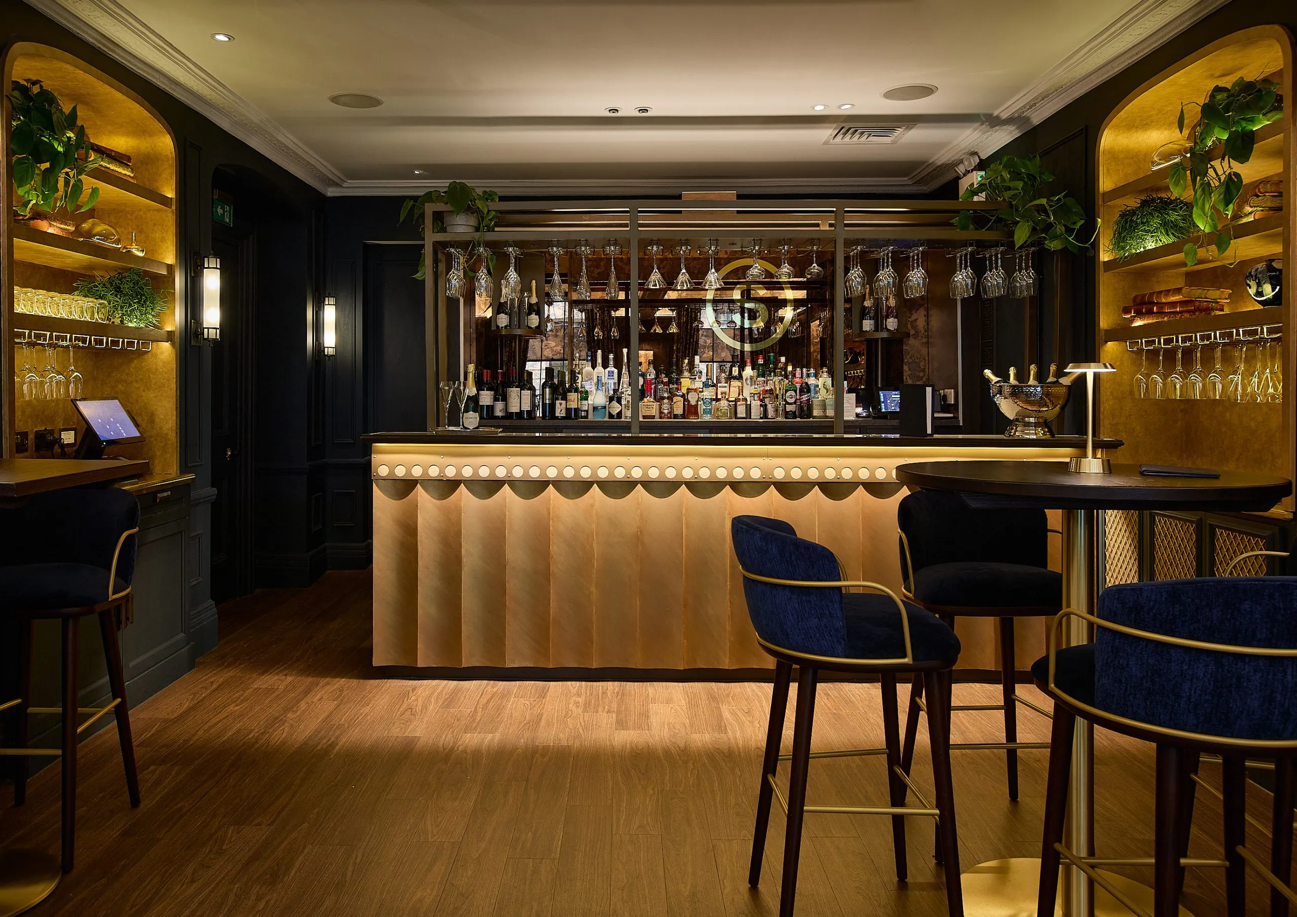 Warm-toned bar interior with scalloped timber counter, navy velvet seating, and brass-lit arched shelving